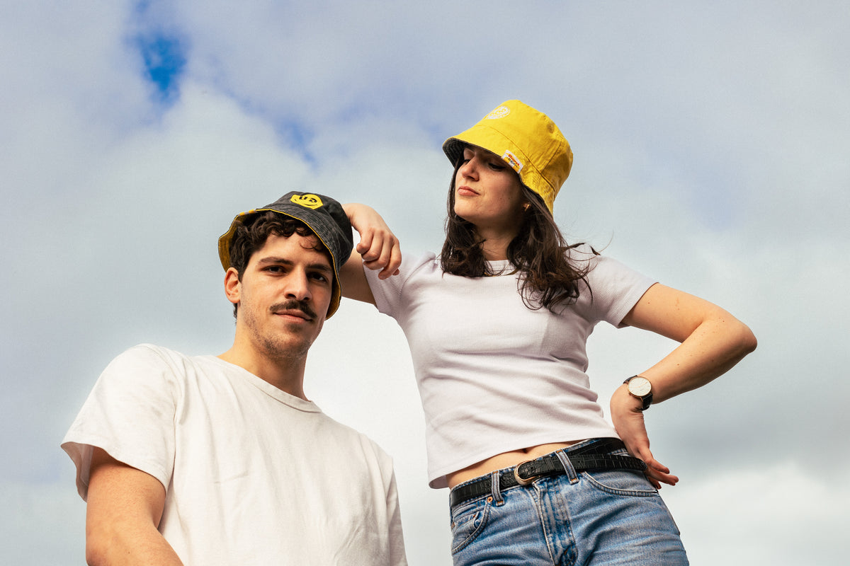 man wearing grey duvel 6,66 bucket hat standing next to woman wearing yellow duvel 6,66 bucket hat