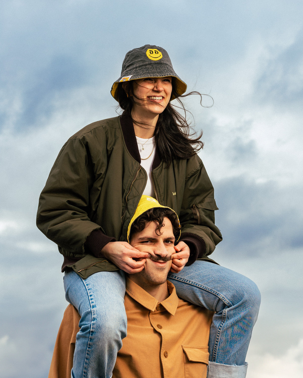 man wearing yellow duvel 6,66 bucket hat with woman wearing grey duvel 6,66 bucket hat sitting on the mans shoulders