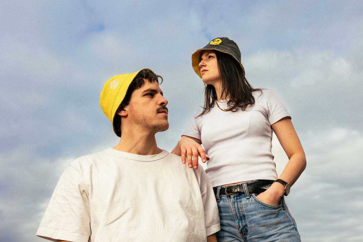 man wearing yellow duvel 6,66 bucket hat standing next to woman wearing grey duvel 6,66 bucket hat